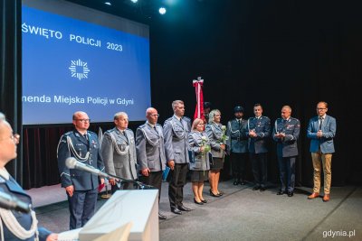 Scena w Konsulacie Kultury w Gdyni, czarne tło, na ekranie napis Święto Policji, na scenie do zdjęcia pozuje kilkoro policjantów i policjantek, komendanci oraz Jakub Ubych