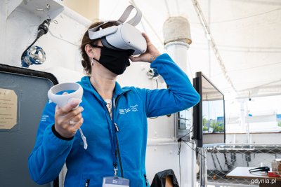 Kobieta w polarze Narodowego Muzeum Morskiego na pokładzie Daru Pomorza. Przykłada do twarzy gogle do wirtualnego zwiedzania, w ręce trzyma sterownik.