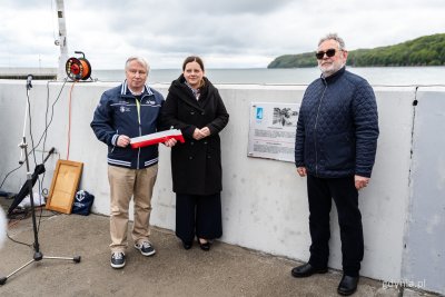 Falochron w marinie Gdynia, tablica pamiątkowa, przed tablicą pozują do zdjęcia: Bogusław Witkowski, który trzyma biało-czerwoną wstęgę, Aleksandra Kosiorek, Janusz Sienkiewicz, w tle zatoka i plaża w Gdyni
