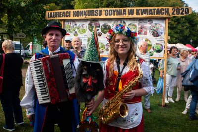 Na tle tablicy ze zdjęciami laureatów konkursu Gdynia w Kwiatach stoją mężczyzna i kobieta w regionalnych, kaszubskich strojach