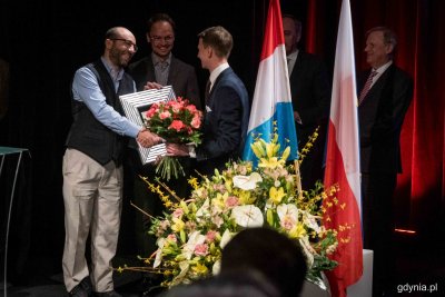 Grupa osób na scenie Konsulatu Kultury w Gdyni. Laureat odbiera nagrody od członków kapituły konkursu, na środku kwiaty oraz flagi: europejska, luksemburska i polska