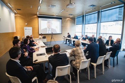 Sala w PPNT Gdynia. Stół ustawiony w literę U, siedzą włodarze miast, gmin i powiatów. Na ekranie wyświetla się połączenie wideo, za oknami fragment Gdyni