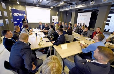Sala konferencyjna w Gdynia Infobox. Stoły na środku, przy stołach kilkadziesiąt osób. Prezentacja wyświetlana na ekranie. Kobieta stoi obok ekranu i przemawia do uczestników spotkania.