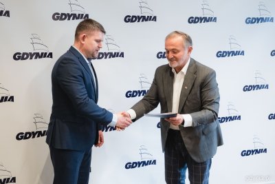 Wojciech Szczurek wręcza teczkę Robertowi Rondudzie, panowie uściskują sobie dłoń. W tle biała ścianka z logotypami Gdyni w kolorze granatowym