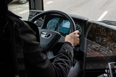 Kabina kierowcy autobusu. Widoczna kierownica, ręce mężczyzny na kierownicy, wyświetlacz, za szybą ulica