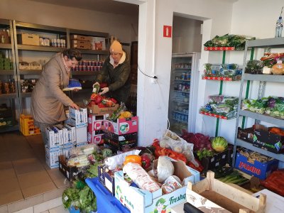 Sklep społeczny w Orłowie przy ul. Lotników. Na zdjęciu pomieszczenie, dwie kobiety układające warzywa na półce, regały z produktami spożywczymi.