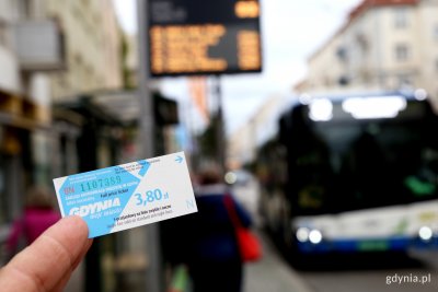 Na pierwszym planie papierowy, jednorazowy bilet ZKM Gdynia. Bilet biało-błękitny z logotypem Gdyni oraz ceną 3,80zł. W drugim planie - rozmazany - trolejbus na przystanku.
