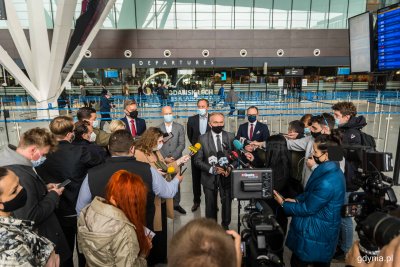 Wnętrze terminala pasażerskiego lotniska. Grupa dziennikarzy, Wojciech Szczurek przy mikrofonach. W tle odprawa.