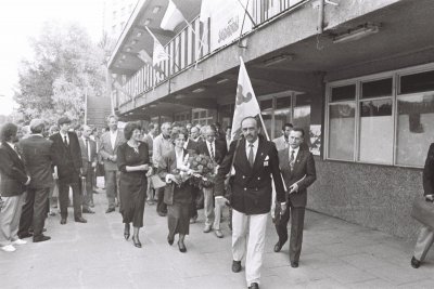 Czarno-białe zdjęcie, na którym widać grupę osób. Z przodu idzie Szymon Pawlicki, w tle widoczne są flagi 