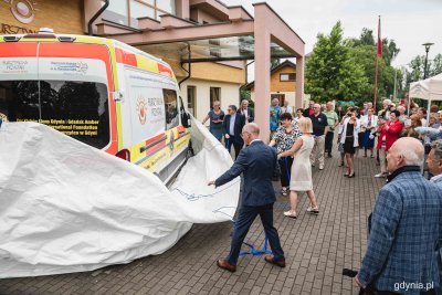 Plac przed hospicjum w Gdyni, tłum ludzi, na środku karetka, trzy osoby ściągają z niej biały pokrowiec, w tle budynek