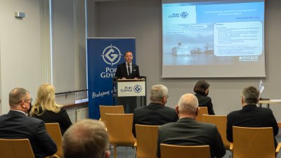 Sala konferencyjna, mówca przy katedrze, obok ekran z wyświetlanym obrazem z projektora. Na sali goście na krzesłach
