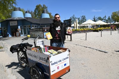 na zdjęciu rower cargo na plaży załadowany książkami, przy rowerze czarnowłosy uśmiechnięty mężczyzna