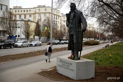Pomnik Józefa Piłsudskiego na terenie zielonym. Obok chodnik i droga rowerowa, aleja Piłsudskiego. Budynki w tle