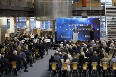 Spotkanie w sali biblioteki Uniwersytetu Gdańskiego. Na środku scena z podwyższeniem, ekran z niebieskim tłem, przy katedrze przemawia Wojciech Szczurek. Przed sceną rzędy krzeseł, na których siedzą goście w formalnych strojach