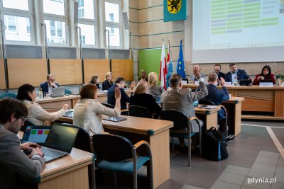 Sala obrad rady miasta, na wyświetlaczu lista nazwisk, kilkanaście osó siedzi w ławach, osoby podnoszą ręce i głosują na uchwały
