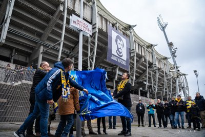 Stadion, tabliczka z nazwą placu, w tle zdjęcie Janusza Kupcewicza, grupa ludzi ściąga materiał i odsłania tablicę