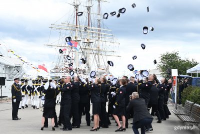 Grupa osób w marynarskich mundurach stoi na nabrzeżu, wyrzucają w górę czapki. W tle Dar Pomorza