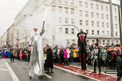 Skrzyżowanie ulic Świętojańskiej i al. m. Piłsudskiego w Gdyni. W tle budynek Urzędu Miasta Gdyni. W głębi ulicy tłum ludzi, na środku postaci odgrywające jedną ze scenek. Jedna przebrana za anioła na szczudłach, druga za diabła.