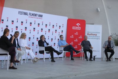 Na amfiteatralnej scenie na krzesłach siedzi 7 osób. Za nimi banery reklamujące festiwal NNW.