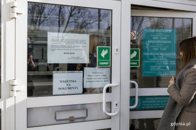 Drzwi wejściowe do budynku Urzędu Miasta Gdyni. Z prawej widać kobietę. Na drzwiach widoczna wrzutnia na dokumenty, zawieszone kartki z informacjami dla interesantów.
