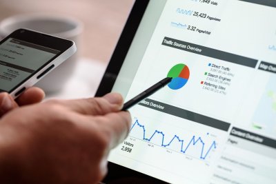 Ekran laptopa z widocznymi wykresami kołowymi i statystykami na stronie internetowej, przed ekranem ręka z długopisem wskazująca na ekran