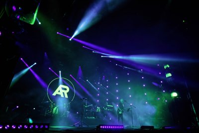 Koncert Artura Rojka w Open'er Park. Widoczne postaci na ciemnej scenie. Scena oświetlona niebieskimi, granatowymi i turkusowymi światłami. Na drugim planie widoczny logotyp AR - występującego Artura Rojka.