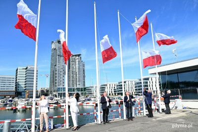 Nabrzeż przy marinie yacht park, kilka osób stoi przy brzegu i wciąga biało-czerwone flagi na maszt. W tle jachty przy kejach, sea towers, hotel marriott