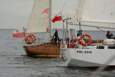 Na zdjęciu dwa jachty na morzu. Z tyłu jacht drewniany, w kolorze brązowym, siedzi na nim mężczyzna. Z przodu jacht w kolorze białym.