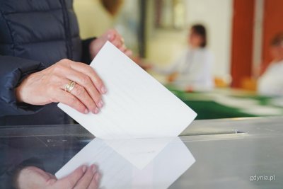 Lokal wyborczy, urna, kobieca dłoń wrzuca do urny białą kartkę