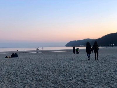 Plaża w Gdyni po zachodzie słońca. Kilka osób siedzi na piasku. Para spaceruje, trzymając się za ręce.