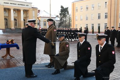 Czterech mężczyzn w mundurach klęczy przy promocji oficerskiej. Mężczyzna dotyka pałaszem lewego ramienia jednego z nich