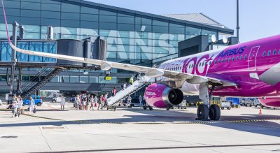 Płyta lotniska w Gdańsku. Ludzie wsiadają do samolotu WizzAir, widoczny rękaw i budynek terminala. Słoneczna pogoda