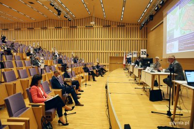 Na zdjęciu utrzymana w drewnie sala konferencyjna Pomorskiego Parku Naukowo-Technologicznego GDYNIA. Po lewej na niektórych siedziskach zasiadają Radni Miasta Gdyni. Po prawej stronie na scenie ustawione stoły, przy których siedzą przewodnicząca RMG wraz ze swoimi zastępcami.