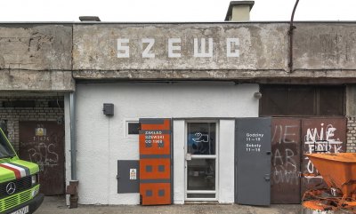 Zdjęcie frontu zakładu szewskiego. Po bokach maszyny. Nad drzwiami zakładu napis 