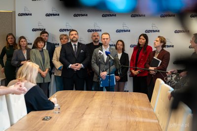 Sala w urzędzie miasta Gdyni, w tle logotypu miasta na białym tle, grupa radnych przemawia do mikrofonu, stół, fragment kamery