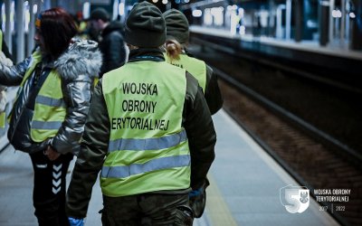 Kobieta w wojskowym mundurze idzie peronem kolejowym. Na plecach zielona kamizelka z napisem 