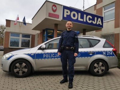 Policjant w mundurze pozuje do zdjęcia. Za nim służbowy radiowóz i budynek komisariatu policji na Oksywiu