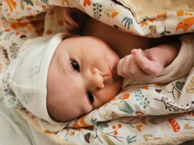 Noworodek o ciemnych oczach zawinięty w biały becik z kolorowymi wzorkami.