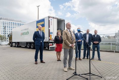 Grupa ludzi na parkingu przy GCS. Prezydent Wojciech Szczurek przemawia do mikrofonów. W tle ciężarówka z naczepą i logo Gdyni na tle ukraińskiej flagi