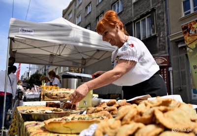 Stragan z jedzeniem obsługiwany przez rudowłosą kobietę. Stragan ustawiony na powietrzu, na ul. Świętojańskiej w Gdyni w ramach festiwalu Kulinarna Świętojańska.