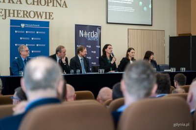 Sala audytoryjna w Wejherowie, na scenie stół prezydialny, siedzi kilka osób, wojewoda pomorskie przemawia przez mikrofon