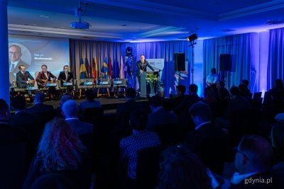 Sala konferencyjna, widownia na krzesłach, na scenie kilkoro gości siedzi w fotelach, mężczyzna przemawia przy mównicy