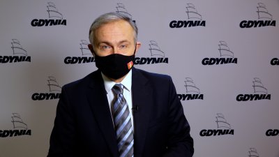 Prezydent Gdyni Wojciech Szczurek ubrany w ciemny garnitur, białą koszulę i krawat w poprzeczne pasy. Ma ubraną czarną maseczkę zasłaniającą usta i nos. Maseczka ozdobiona herbem Gdyni. W tle ścianka medialna w kolorze białym i logotypami Gdyni.