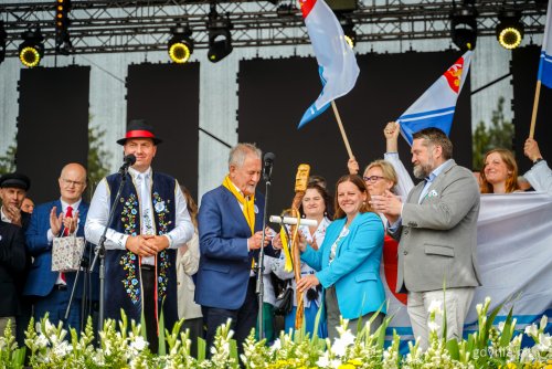 Scena na festynie, grupa osób - samorządowców, przedstawicieli rządu, radnych, na środku Ryszard Kalkowski, Jan Wyrowiński, Aleksandra Kosiorek trzymająca klekę - laskę sołecką, obok Tadeusz Szemiot, widoczne flagi Gdyni