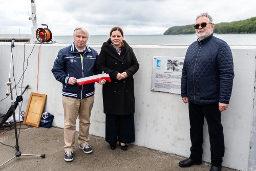 Falochron w marinie Gdynia, tablica pamiątkowa, przed tablicą pozują do zdjęcia: Bogusław Witkowski, który trzyma biało-czerwoną wstęgę, Aleksandra Kosiorek, Janusz Sienkiewicz, w tle zatoka i plaża w Gdyni