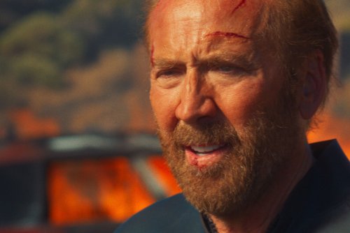Kadr z filmu „Surfer”. Poraniony bohater stoi na tle płonącego samochodu.