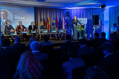 Sala konferencyjna, widownia na krzesłach, na scenie kilkoro gości siedzi w fotelach, mężczyzna przemawia przy mównicy