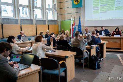 Sala obrad rady miasta, na wyświetlaczu lista nazwisk, kilkanaście osó siedzi w ławach, osoby podnoszą ręce i głosują na uchwały