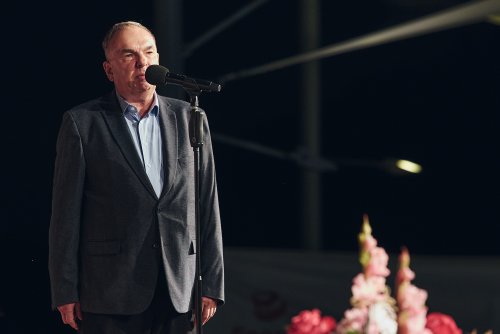 Meżczyzna w ciemnym garniturze na scenie, obok kwiaty
