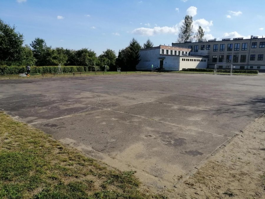 Na zdjęciu asfaltowe boisko, popękana nawierzchnia, w tle budynek szkoły i drzewa.